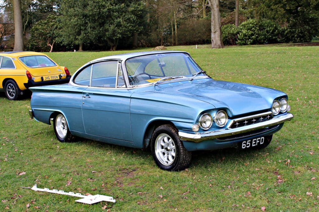Gracias por la corrección. Aquí tienes: La imagen muestra un Ford Capri de 1965 azul estacionado en una zona de césped, con otro coche amarillo en el fondo. El coche azul tiene un diseño elegante y vintage, con una parrilla frontal distintiva y faros dobles a cada lado. La matrícula del coche azul dice "65 EFD". El coche amarillo en el fondo tiene una matrícula que dice "JHG 990M". El entorno parece ser un parque o un área abierta con árboles y césped. También hay un objeto blanco en el césped frente al coche azul, que parece ser una pieza o accesorio de coche. Esta imagen es interesante porque muestra coches clásicos, que a menudo son apreciados por su diseño y valor histórico.