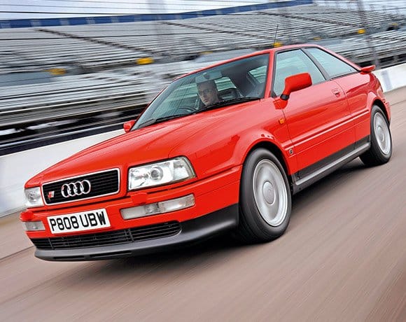 La imagen muestra un Audi 80 Competition Quattro rojo conduciendo en una pista de carreras. El coche tiene una matrícula con el número "P808 UBW" y presenta el logotipo de Audi en la parrilla frontal. El coche parece estar en movimiento, sugiriendo que está siendo conducido a alta velocidad. El fondo incluye gradas desenfocadas, indicando que el coche está en un circuito de carreras profesional. Esta imagen es interesante porque captura la naturaleza dinámica y deportiva del vehículo, destacando sus capacidades de rendimiento en una pista de carreras.