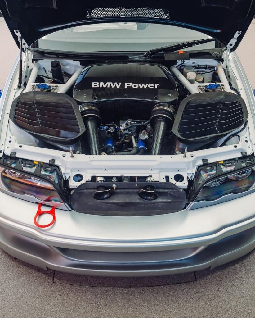La imagen muestra el compartimento del motor de un BMW M3 GTR con el capó abierto. La cubierta del motor muestra prominentemente el texto "BMW Power". El compartimento del motor está limpio y bien organizado, con componentes visibles como los sistemas de admisión de aire a ambos lados, varias mangueras y elementos estructurales. Este coche es famoso por su aparición en el videojuego "Need for Speed: Most Wanted", donde juega un papel central. La imagen es interesante y relevante para los entusiastas del automovilismo y los fans del juego.