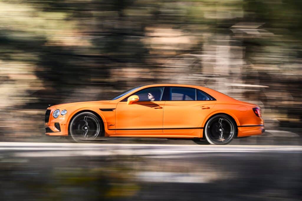 La imagen muestra un Bentley Flying Spur Speed 2025 de color naranja conduciendo a alta velocidad por una carretera. El fondo está desenfocado para enfatizar el movimiento. El coche destaca por su diseño elegante y deportivo, con el logotipo distintivo de Bentley en las ruedas. La imagen captura la esencia del lujo y el rendimiento en el diseño automotriz, resaltando las características deportivas y elegantes del vehículo.
