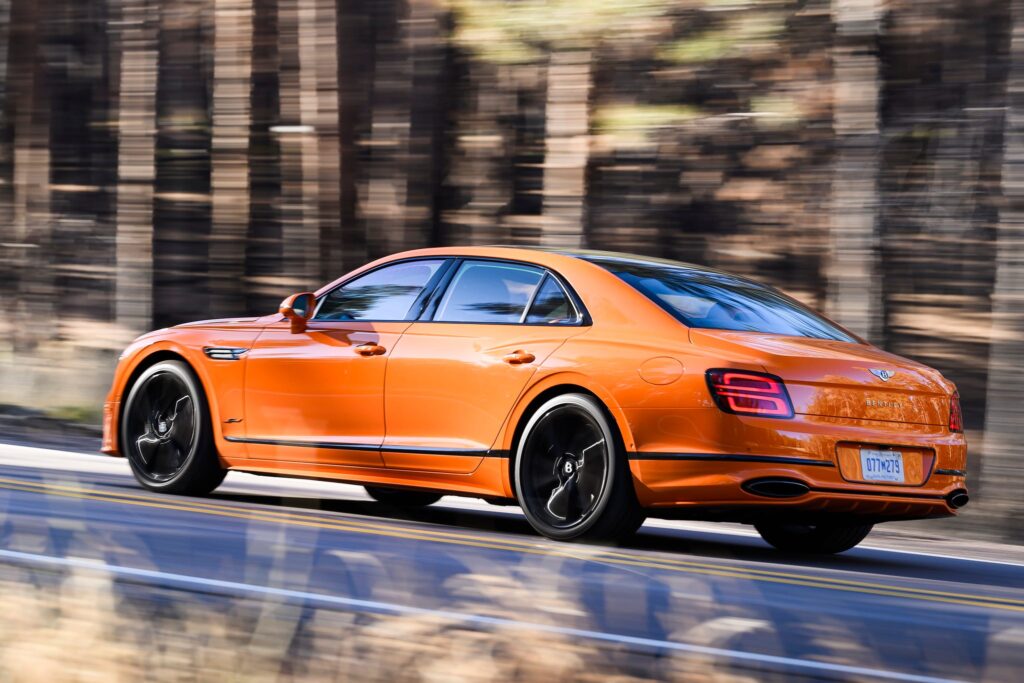 La imagen muestra un Bentley Flying Spur Speed 2025 de color naranja conduciendo por una carretera con un fondo de bosque desenfocado, lo que indica movimiento. El coche tiene ruedas negras con un diseño distintivo y una carrocería aerodinámica y elegante. La matrícula dice "Q7M 279". Esta imagen es interesante porque captura la elegancia y el rendimiento de un coche de lujo en movimiento, destacando su diseño y la experiencia dinámica de conducción que ofrece.
