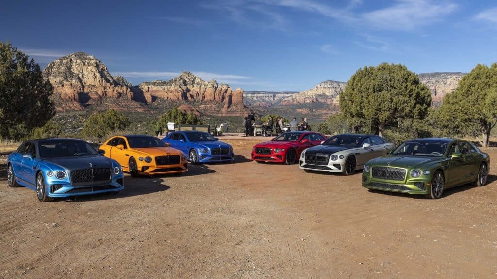 La imagen muestra una alineación de seis Bentley Flying Spur Speed 2025 en diferentes colores. Los coches están estacionados en una zona de tierra con un fondo escénico de formaciones rocosas rojas y árboles, lo que sugiere una ubicación en una región desértica o montañosa. Los colores de los coches incluyen azul, naranja, otro tono de azul, rojo, plateado y verde. Hay algunas personas en el fondo, posiblemente involucradas en un evento de coches o una sesión de fotos. La imagen es interesante porque muestra la variedad de colores disponibles para este modelo de coche en particular y destaca los coches en un entorno pintoresco.