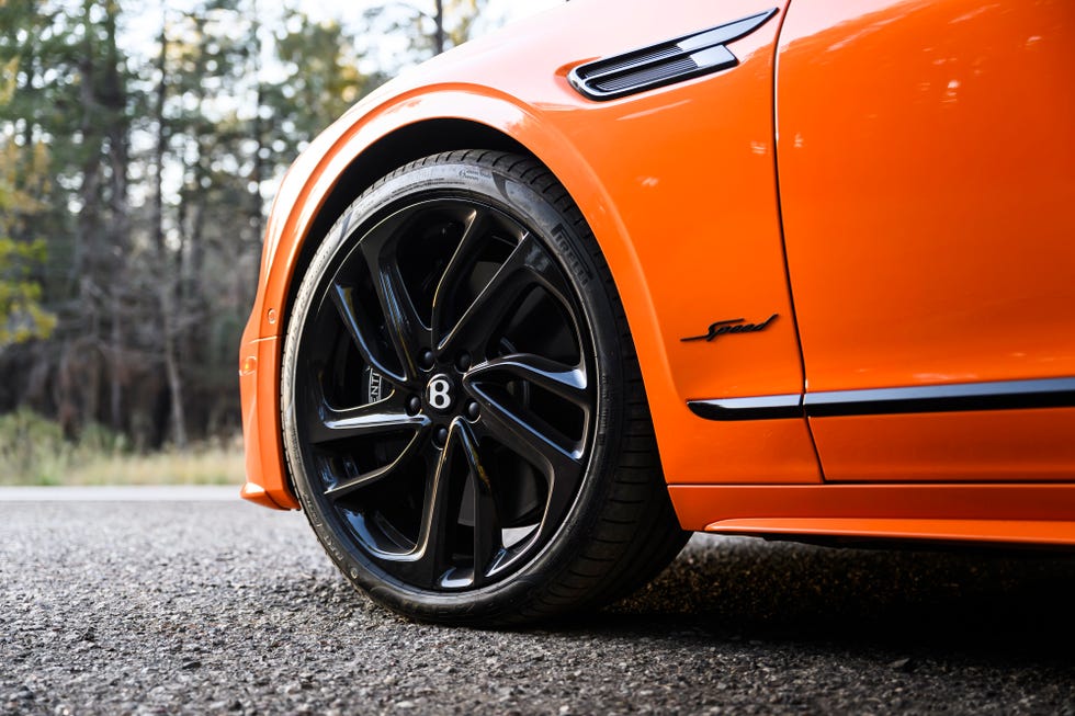 La imagen muestra un primer plano de la rueda delantera y parte del lateral de un Bentley Flying Spur Speed 2025 de color naranja. La rueda tiene un diseño de llanta de aleación negra con el logotipo de Bentley en el centro. En el lateral del coche, se puede ver una insignia que dice "Speed", indicando que es un modelo de alto rendimiento. La marca del neumático "Pirelli" es visible en el flanco. Esta imagen destaca los aspectos de lujo y rendimiento del Bentley, mostrando los elementos de diseño y marca característicos de los vehículos de alta gama.