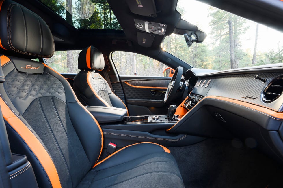 "La imagen muestra el interior de un Bentley Flying Spur Speed 2025. Los asientos están tapizados en cuero negro con detalles y costuras en naranja, presentando un patrón acolchado en los respaldos. Los reposacabezas tienen bordada la palabra 'Speed'. El tablero y la consola central están diseñados con materiales de alta calidad, incluyendo fibra de carbono y cuero, también con detalles en naranja. El coche tiene un techo solar panorámico, y se pueden ver el volante y varios controles. El diseño general sugiere un vehículo de alto rendimiento y premium.