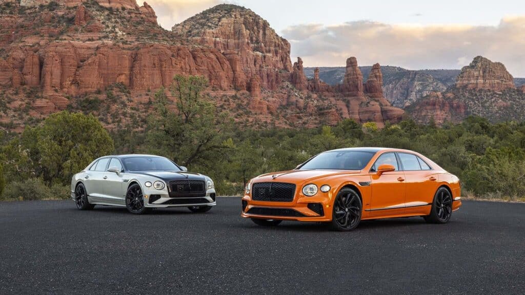 La imagen muestra dos coches de lujo estacionados sobre una superficie pavimentada con un fondo escénico de formaciones rocosas rojas y vegetación. El coche de la izquierda es un Bentley Flying Spur Speed 2025 de dos tonos, con una carrocería blanca y detalles en negro. El coche de la derecha es un Bentley Flying Spur Speed 2025 de color naranja brillante con detalles en negro. Ambos coches tienen un diseño elegante y moderno, destacando como modelos de alta gama de un fabricante de automóviles premium. El entorno parece ser una región desértica o montañosa, lo que añade atractivo visual a la imagen al resaltar tanto el diseño de los coches como la belleza del paisaje natural.