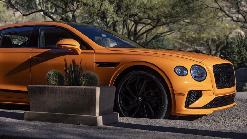 La imagen muestra un primer plano de un Bentley Flying Spur Speed 2025 de color naranja. La imagen se enfoca en la parte delantera y lateral del vehículo, destacando su diseño elegante con acentos negros, incluyendo la parrilla, las ruedas y las ventilaciones laterales. El fondo incluye un entorno natural con árboles y plantas, y en primer plano hay una jardinera de concreto con cactus. El diseño distintivo y el color vibrante del coche lo hacen destacar, resaltando su apariencia de alta gama y estilo.