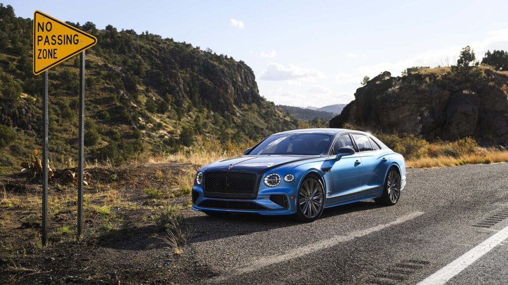 La imagen muestra un Bentley azul estacionado al lado de una carretera en una zona montañosa. A la izquierda de la imagen, hay una señal de tráfico triangular amarilla que dice 'NO PASSING ZONE'. El fondo presenta colinas rocosas con vegetación escasa, lo que indica un entorno pintoresco y posiblemente remoto. El diseño elegante del coche y el entorno escénico hacen que la imagen sea visualmente atractiva y relevante para mostrar la elegancia y el rendimiento del coche en un entorno natural.