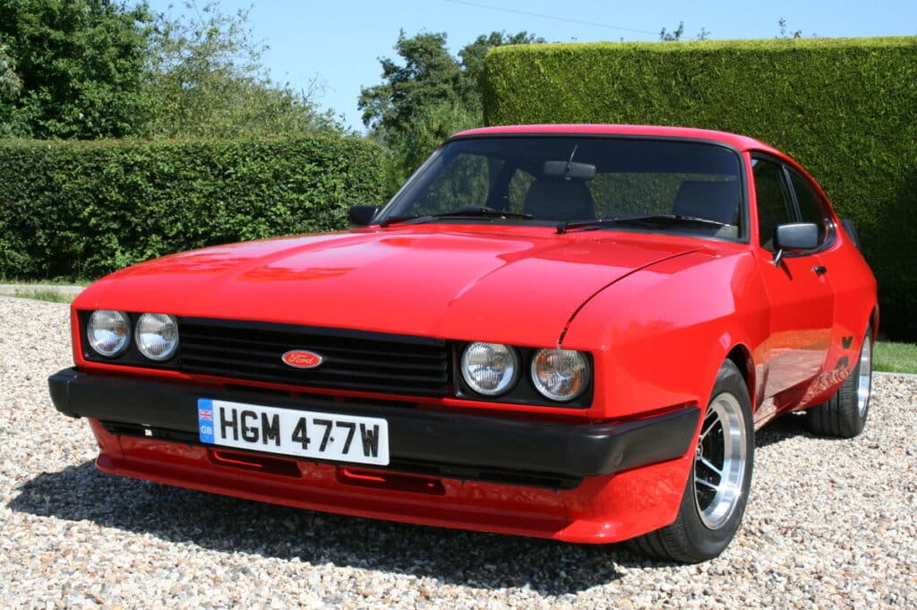 Un coche clásico Ford Capri rojo estacionado en una superficie de grava, con matrícula HGM 477W. El coche tiene un diseño aerodinámico con faros redondos dobles y un parachoques negro. Al fondo, hay un seto verde y árboles, lo que sugiere que el coche está en un entorno rural o de jardín.
