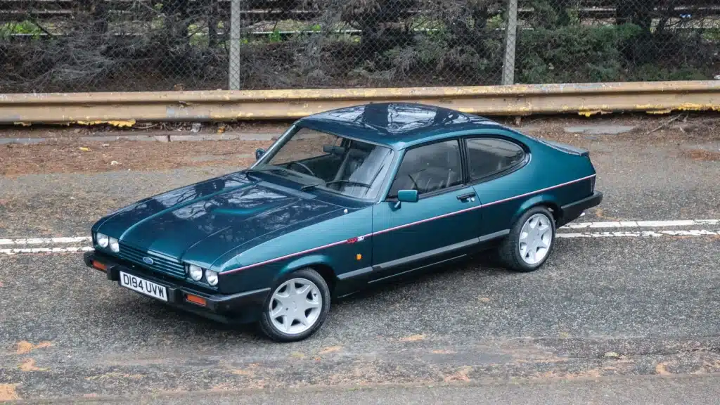 La imagen muestra un Ford Capri Brooklands, un coche clásico conocido por su diseño distintivo y su importancia histórica. El coche está estacionado en una carretera con una barandilla metálica y una valla de alambre en el fondo. El Ford Capri Brooklands es notable por su apariencia elegante y deportiva, caracterizada por su color verde oscuro, forma aerodinámica y llantas de aleación estilizadas. La matrícula dice "D194 UVW". Este modelo es particularmente interesante para los entusiastas de los coches debido a su producción limitada y su estatus icónico en la historia automotriz.