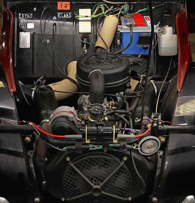 La imagen muestra el compartimento del motor de un Citroën 2CV Charleston. El motor está ubicado en el centro con varios componentes y cables visibles. Elementos notables incluyen un ventilador grande en la parte inferior, una batería en el lado derecho y varias mangueras y cables conectados a diferentes partes del motor. La imagen es interesante porque proporciona una vista detallada de la disposición y los componentes del motor, lo cual puede ser útil para entender la mecánica o para propósitos de restauración.