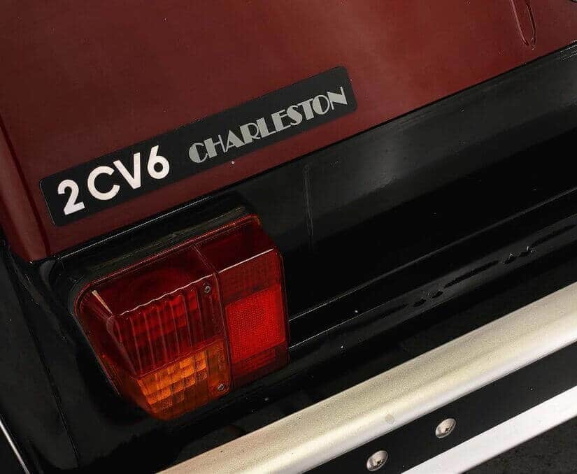 La imagen muestra la parte trasera de un coche, enfocándose específicamente en la luz trasera izquierda y una insignia en el maletero. La insignia dice "2CV6 CHARLESTON". La luz trasera tiene una lente roja y ámbar. Esta imagen es interesante porque destaca un modelo específico del Citroën 2CV, conocido como el "Charleston", que es una edición especial de este icónico coche francés.