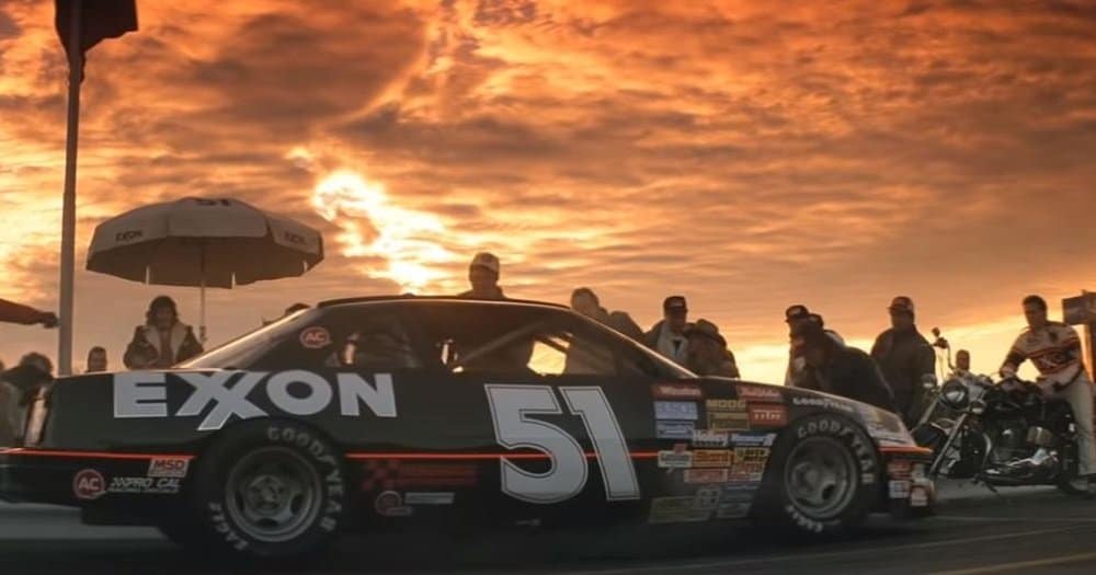 Un coche de carreras negro con el número 51 y el logotipo de Exxon, estacionado y rodeado por un equipo de boxes y equipo. Al fondo, un cielo dramático al atardecer con tonos naranjas y amarillos.