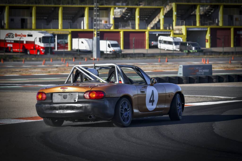 La imagen muestra un coche de carreras en una pista. El coche es un convertible con una jaula antivuelco instalada y tiene el número 4 en su costado. El coche parece estar en movimiento, como lo indica el ligero desenfoque de las llantas y el ángulo del coche en la pista. En el fondo, hay varios vehículos, incluyendo un gran camión rojo y blanco con el logotipo "JEG" y otros vehículos de apoyo. El escenario parece ser un circuito de carreras, con barreras, conos y un cartel que dice "CIRCUIT RICARDO" visible. Esta imagen es interesante porque captura el entorno dinámico de un evento de carreras, mostrando el coche en acción y la infraestructura de apoyo.