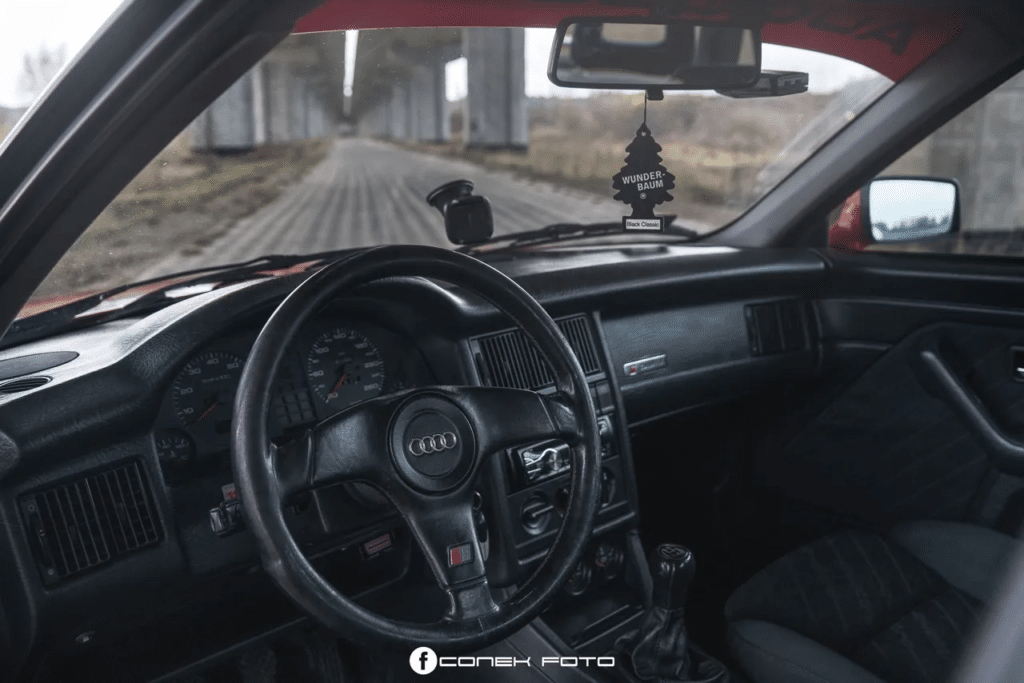 La imagen muestra el interior de un Audi 80 Competition. El área del asiento del conductor y el tablero son claramente visibles. El volante está en el centro, con un logotipo en el medio. El tablero incluye varios indicadores y controles, como un velocímetro y un indicador de combustible. La palanca de cambios está ubicada entre los dos asientos delanteros. Los asientos están tapizados en tela marrón, y los paneles de las puertas también son visibles, con manijas y manivelas para las ventanas. El diseño general parece ser de un vehículo de modelo antiguo, posiblemente de finales del siglo XX. Esta imagen es interesante porque proporciona una visión del diseño y la disposición de los interiores de coches de una era pasada, destacando la simplicidad y funcionalidad de los controles y características.