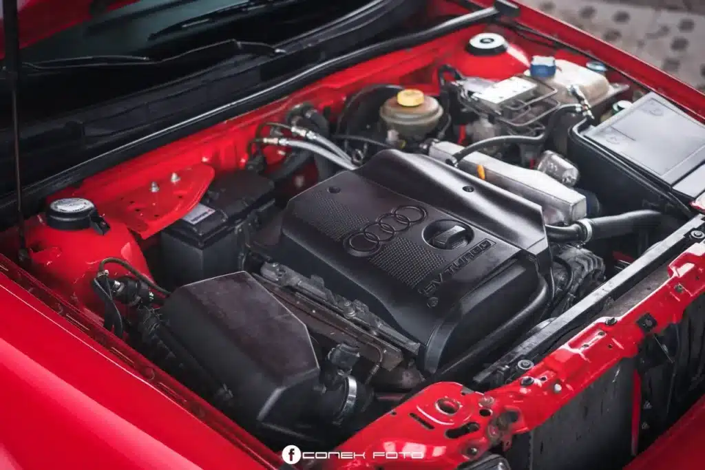 Gracias por la corrección. Aquí tienes: La imagen muestra el compartimento del motor de un Audi 80 Competition Quattro. La cubierta del motor exhibe de manera prominente el logotipo de Audi y el texto "5V TURBO", indicando que se trata de un motor turboalimentado con una configuración de cinco válvulas. El compartimento del motor está limpio y bien organizado, con varios componentes visibles, como la batería, el depósito de refrigerante y el sistema de admisión de aire. Esta imagen es interesante y relevante porque destaca el tipo específico de motor y su configuración, lo cual puede ser de interés para los entusiastas de los coches o aquellos que buscan entender más sobre las capacidades de rendimiento de este vehículo en particular.
