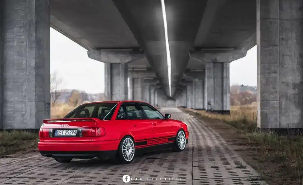 La imagen muestra un Audi 80 Competition Quattro rojo estacionado bajo un gran puente de concreto. El coche está posicionado en un camino pavimentado con césped a ambos lados. La estructura del puente está sostenida por grandes pilares de concreto, y la perspectiva de la imagen crea una sensación de profundidad, atrayendo la mirada del espectador hacia el horizonte. El coche tiene una matrícula con el número "GST 25294" y presenta un alerón trasero y elementos de diseño deportivo. El entorno parece ser una zona industrial o semi-rural, lo que añade contraste entre el coche elegante y el entorno rústico.