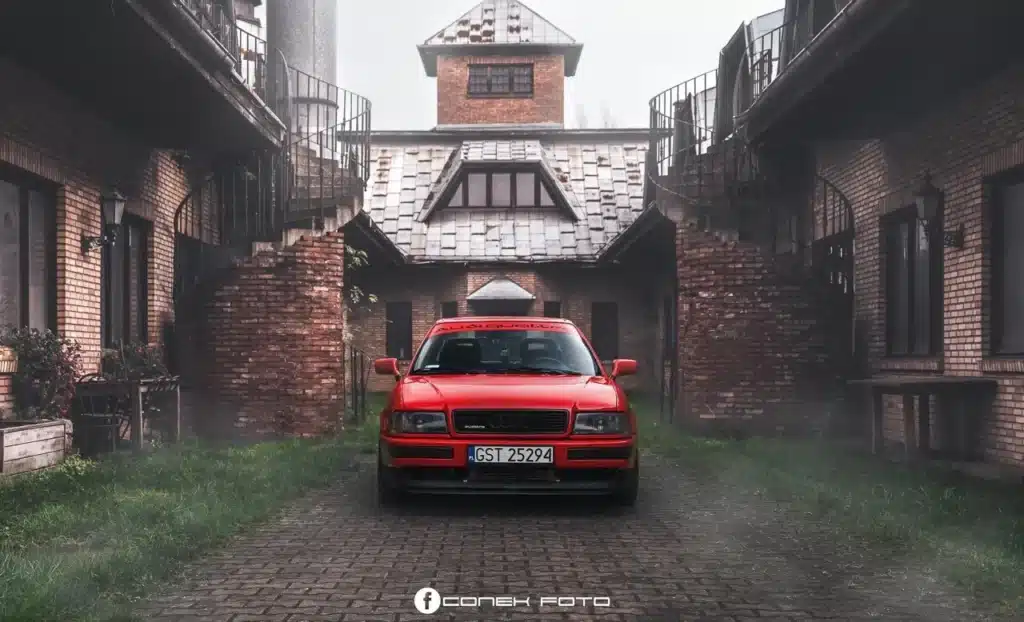 Claro, aquí tienes: La imagen muestra un Audi 80 Competition Quattro rojo estacionado en el centro de un camino de adoquines entre dos edificios de ladrillo. Los edificios tienen una apariencia rústica con escaleras de caracol y balcones a ambos lados. La matrícula del coche dice "GST 25294", y hay un logotipo y texto en la parte inferior de la imagen que dice "CONEK FOTO". El entorno parece ser brumoso o nebuloso, añadiendo una cualidad misteriosa y atmosférica a la escena. La imagen es interesante debido al contraste entre el vibrante coche rojo y los alrededores envejecidos y apagados, creando una composición visualmente impactante.