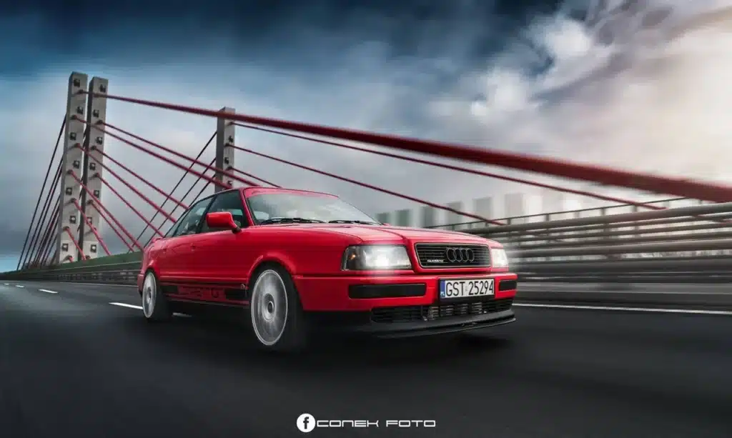 La imagen muestra un Audi 80 Competition Quattro rojo conduciendo sobre un puente. El puente tiene un diseño moderno con cables rojos y pilares de concreto. La matrícula del coche dice "GST 25294". El fondo presenta un cielo nublado, y el coche parece estar en movimiento, creando una escena dinámica y atractiva. La imagen es interesante debido a la combinación del coche elegante y los elementos arquitectónicos del puente, destacando tanto el diseño automotriz como el estructural. El texto "CONEK FOTO" es visible en la parte inferior de la imagen.