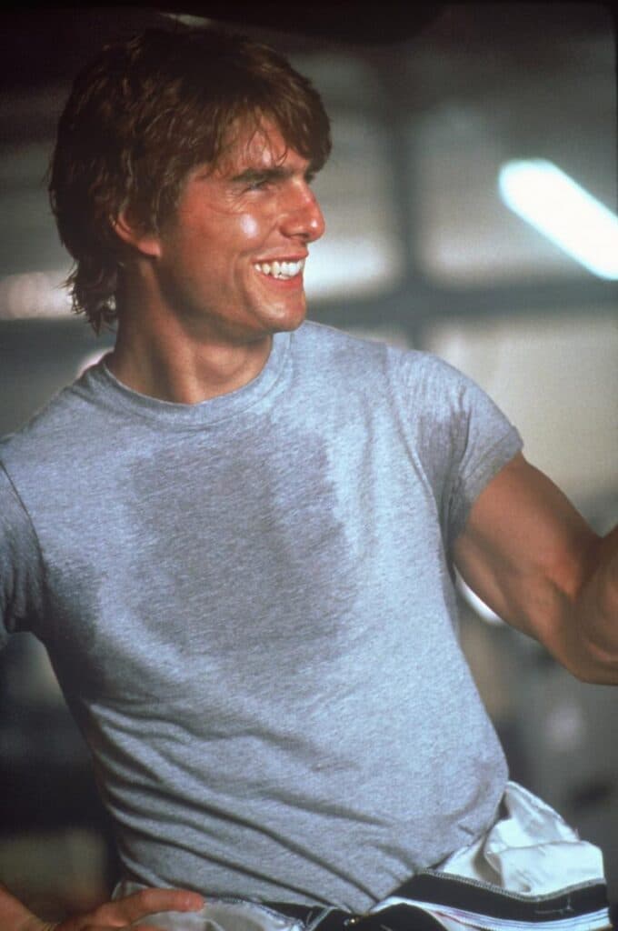 Tom Cruise de frente, mirando hacia su izquierda, con una camiseta gris con manchas oscuras de sudor y jeans azules.