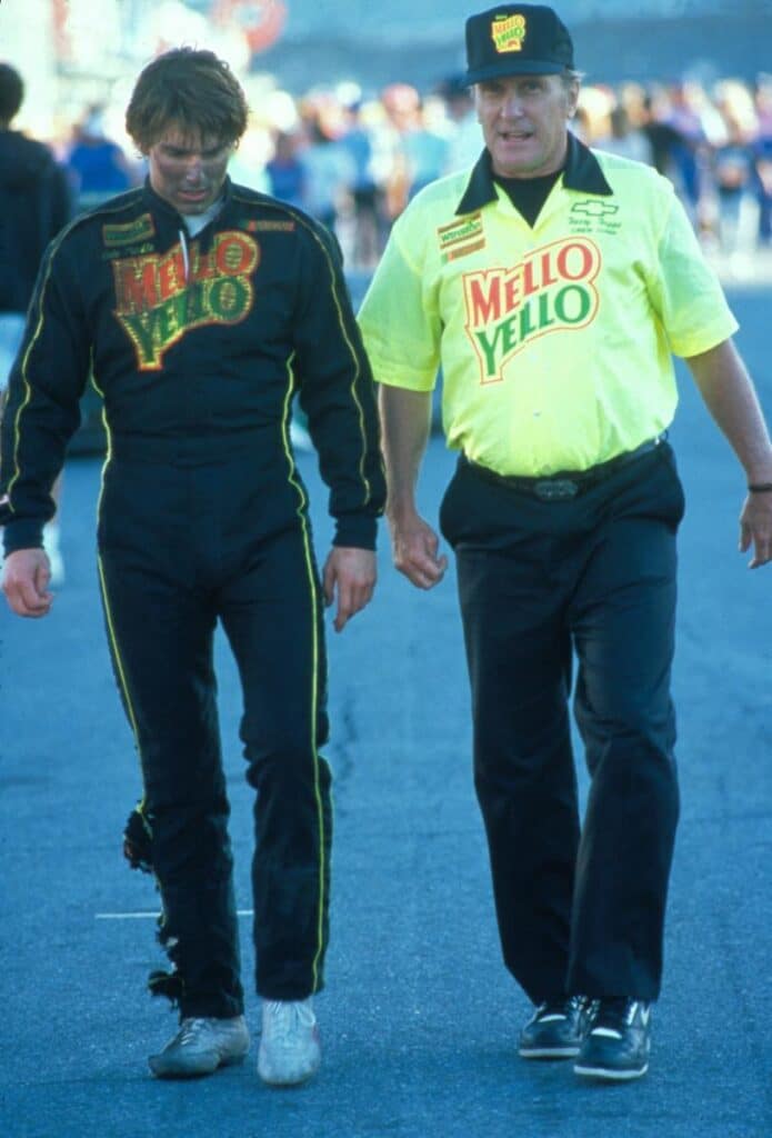 Escena de la película ‘Días de Trueno’ con Tom Cruise como Cole Trickle y Robert Duvall como su mentor Harry Hogge, ambos de pie junto a un coche de carreras en un entorno de pista. El fondo muestra un cielo al atardecer.