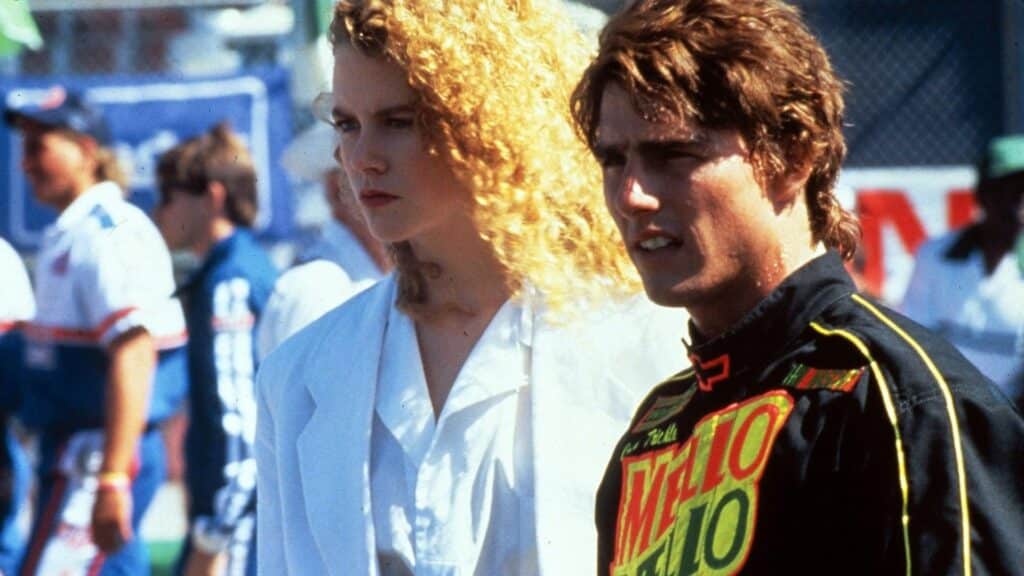 Escena de la película ‘Días de Trueno’ con Nicole Kidman en una camisa blanca y Tom Cruise en un traje de carreras negro con acentos verdes y rojos y la palabra ‘MALTO’ visible en el pecho. Al fondo, un cielo al atardecer y una barrera de seguridad.