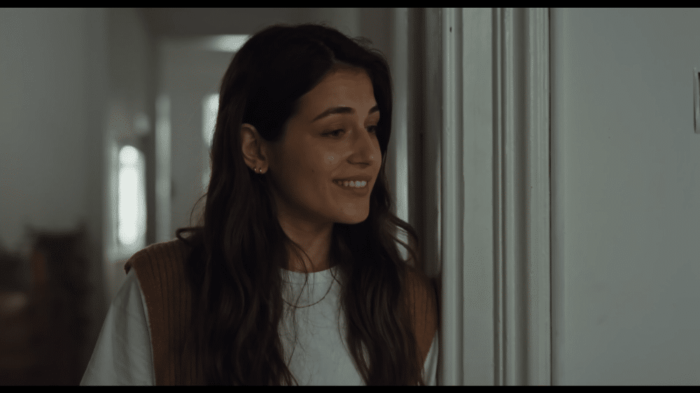 Volvo EX90: La imagen muestra a una mujer de pie en un pasillo, apoyada en el marco de una puerta. Lleva una camisa blanca con un chaleco marrón encima. El pasillo parece ser parte de una casa residencial, con puertas y una ventana visibles en el fondo. La escena captura un momento de quietud y contemplación en un entorno doméstico.