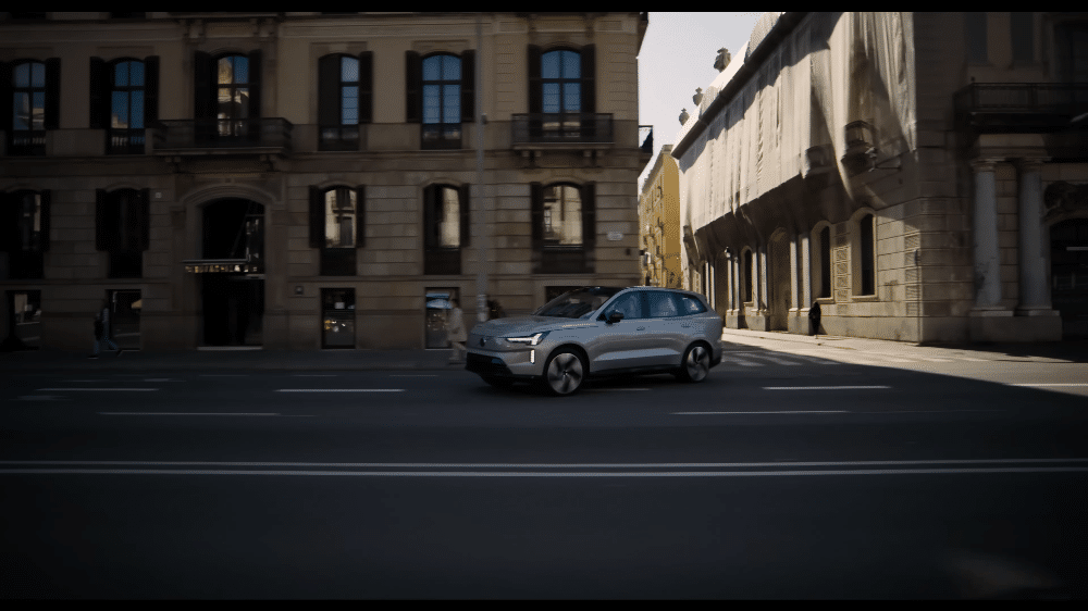 Un Volvo EX90 plateado circula por una calle urbana europea. El vehículo está fotografiado desde un ángulo lateral, con un fondo de edificios clásicos con grandes ventanales. La escena está bañada en luz natural, creando un contraste entre las sombras de los edificios y la iluminación directa sobre el coche.
