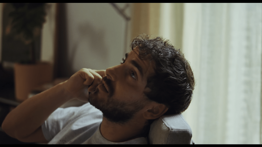 La imagen muestra a un hombre sentado en un sofá, sosteniendo un teléfono en su oído. El rostro del hombre está desenfocado, lo que hace imposible identificarlo. En el fondo, hay una planta en maceta y una cortina. El entorno parece ser un salón, posiblemente en una casa. La imagen captura un momento de comunicación, posiblemente una llamada telefónica, en un ambiente casual y relajado.