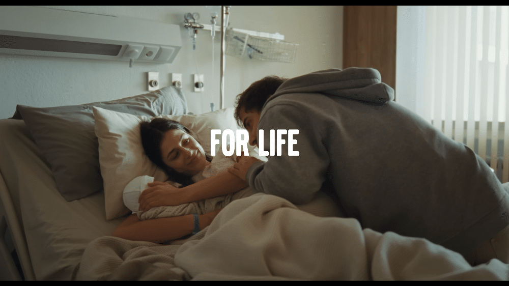 La imagen muestra a una mujer en una cama de hospital, sosteniendo a su recién nacida en brazos. La mujer sonríe con alivio y felicidad, mientras el padre, con una expresión de alegría, está a su lado. El entorno es claramente una habitación de hospital, con equipo médico visible. Esta escena representa el momento de alegría y alivio después de que el sistema de detección de peatones del Volvo evitara un accidente, permitiendo que la madre y su hija estén a salvo