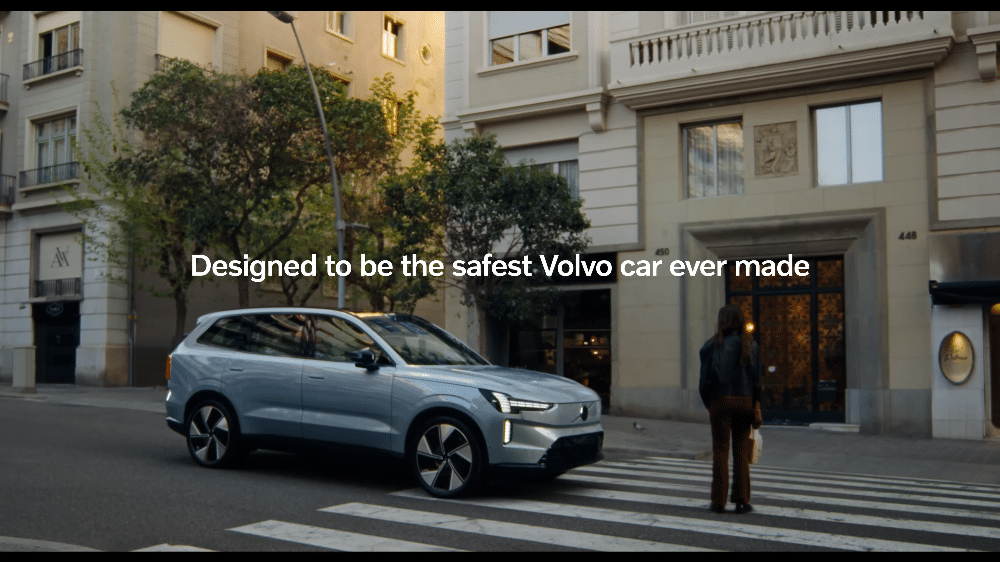La imagen muestra a una mujer embarazada de pie en un paso de peatones, mirando hacia un coche Volvo detenido. El coche ha frenado de repente, evitando un posible accidente. Esta escena destaca la eficacia del sistema de detección de peatones del Volvo, que ha protegido a la futura mamá.
