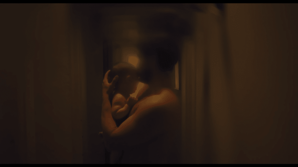 Captura del anuncio de Volvo EX90 en tonos cálidos y oscuros. A través de una puerta entreabierta, se observa a un padre abrazando tiernamente a su bebé en una habitación tenuemente iluminada. La imagen, bañada en tonos dorados y sombras suaves, captura un momento íntimo de conexión paternal.