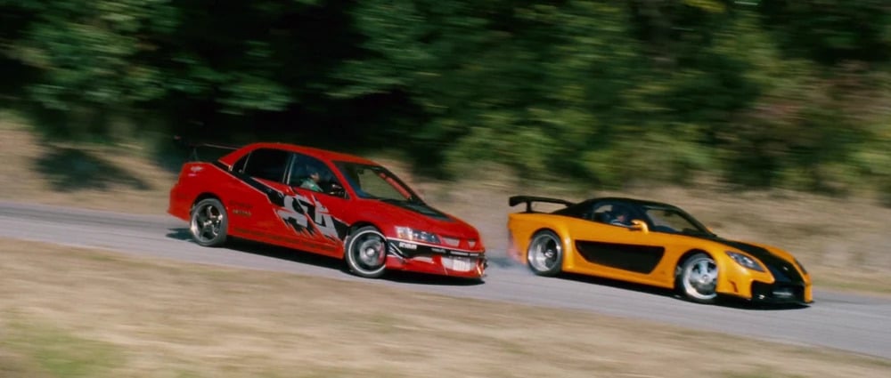La imagen muestra dos coches derrapando en una curva en una carretera. El coche de la izquierda es un Mitsubishi Lancer Evolution rojo con un gran alerón trasero y calcomanías de carreras. El coche de la derecha es un Mazda RX-7 naranja con detalles en negro y un alerón trasero. Ambos coches parecen estar en un derrape controlado, lo que indica una conducción a alta velocidad y técnicas de conducción avanzadas. Esta imagen es interesante y relevante ya que captura la emoción y la habilidad involucradas en el drifting, una actividad popular en el automovilismo y la cultura del automóvil.