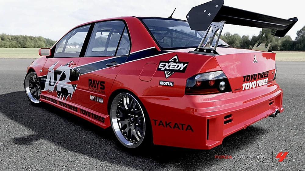 La imagen muestra un Mitsubishi Lancer Evolution IX (EVO 9) rojo modificado con varias calcomanías de carreras y un gran alerón trasero. El coche está estacionado en una superficie pavimentada con un fondo de césped. Las calcomanías incluyen marcas como Toyo Tires, Exedy, Rays Engineering, Optima, Takata y Motul. El coche parece estar preparado para carreras, lo que se indica por la jaula antivuelco visible a través de las ventanas y las modificaciones aerodinámicas.