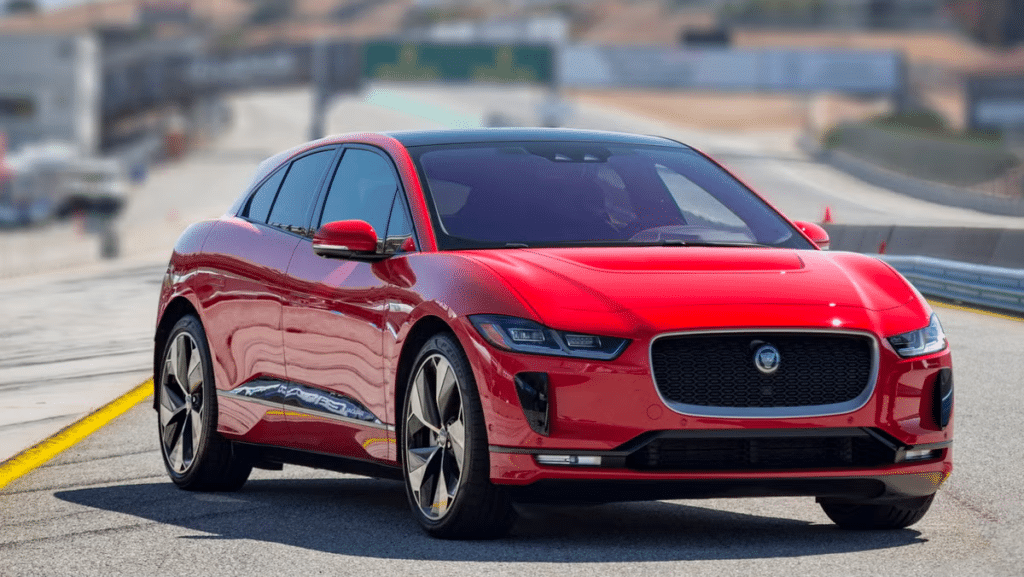La imagen muestra un Jaguar I-Pace rojo, un SUV eléctrico, en una pista de carreras. El coche está posicionado en una carretera pavimentada con un fondo borroso, lo que indica que podría estar en movimiento o en una prueba de conducción. El diseño elegante, la parrilla frontal distintiva y los faros modernos hacen que el vehículo destaque. Esta imagen es interesante y relevante ya que muestra un vehículo eléctrico de lujo, destacando los avances en tecnología automotriz y la transición hacia el transporte sostenible.