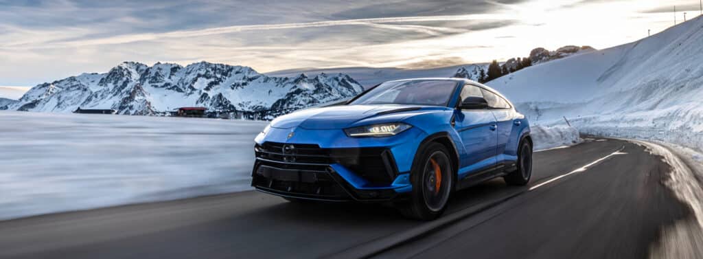 "La imagen muestra un Lamborghini Urus S azul conduciendo por una carretera de montaña cubierta de nieve. El fondo presenta montañas nevadas bajo un cielo parcialmente nublado. El vehículo parece estar en movimiento, como lo indica la nieve borrosa en primer plano y la postura dinámica del coche. Esta imagen es interesante y relevante porque muestra el rendimiento y la capacidad del vehículo en un entorno invernal desafiante, destacando su diseño y la ingeniería adecuada para tales condiciones.