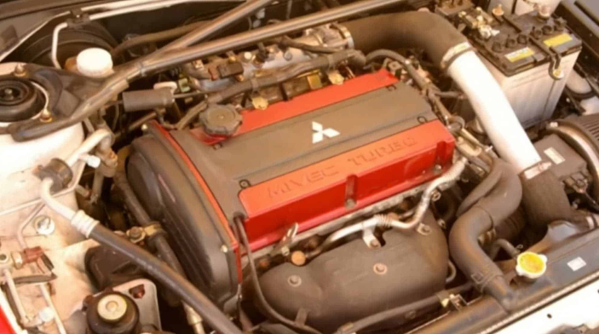 La imagen muestra el compartimento del motor de un Mitsubishi Lancer Evolution IX (EVO 9). El motor es un 4G63T, un motor turboalimentado de 2.0 litros y cuatro cilindros en línea. La tapa de válvulas roja con el logotipo de Mitsubishi y la inscripción "MIVEC TURBO" es una característica distintiva de este motor. El compartimento del motor también incluye varios componentes como la batería, la admisión de aire y diversos tubos y cables. Este motor es conocido por su rendimiento y es popular entre los entusiastas de los coches por su potencial de ajuste y fiabilidad.