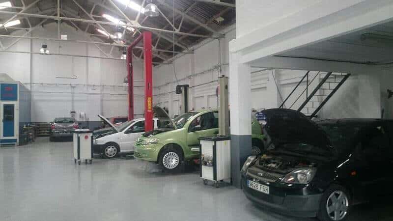 La imagen muestra el interior de un taller de reparación de automóviles. Hay varios coches en el taller, cada uno posicionado en diferentes estaciones de trabajo. Algunos coches tienen el capó abierto, lo que indica que están siendo sometidos a mantenimiento o reparación. El taller está equipado con diversas herramientas y equipos, incluyendo un elevador hidráulico rojo, carros de herramientas y máquinas de diagnóstico. El techo es alto con iluminación industrial, y hay una escalera que conduce a un nivel superior. El entorno parece organizado y bien iluminado, adecuado para trabajos mecánicos.