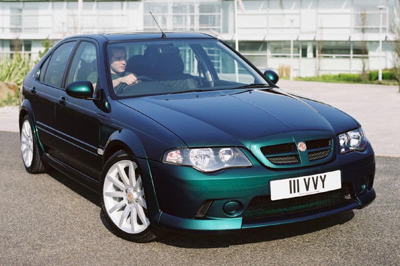 "La imagen muestra un MG ZS 180 de 2004, un sedán deportivo compacto. El coche está estacionado al aire libre frente a un edificio moderno. Presenta una pintura metálica verde oscuro con un acabado brillante, llantas de aleación blancas y una parrilla frontal distintiva con el emblema de MG. La matrícula dice 'III VVY'. Este coche es interesante debido a su diseño deportivo y al hecho de que representa un modelo orientado al rendimiento de la marca MG, conocida por su herencia automovilística británica.