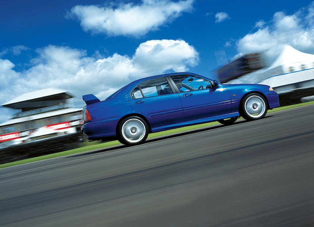 La imagen muestra un coche MG ZS 180 azul conduciendo a alta velocidad en una pista de carreras. El fondo está borroso, indicando movimiento, y hay estructuras como una tribuna y carpas visibles, sugiriendo un evento de carreras o una prueba de manejo. El coche parece estar en movimiento, enfatizando sus capacidades de velocidad y rendimiento. El cielo azul claro con nubes dispersas añade una sensación dinámica y vibrante a la imagen. Esta imagen es interesante y relevante ya que destaca el rendimiento del coche en un entorno de carreras, lo cual puede ser atractivo para los entusiastas de los coches y posibles compradores interesados en vehículos de alta velocidad.