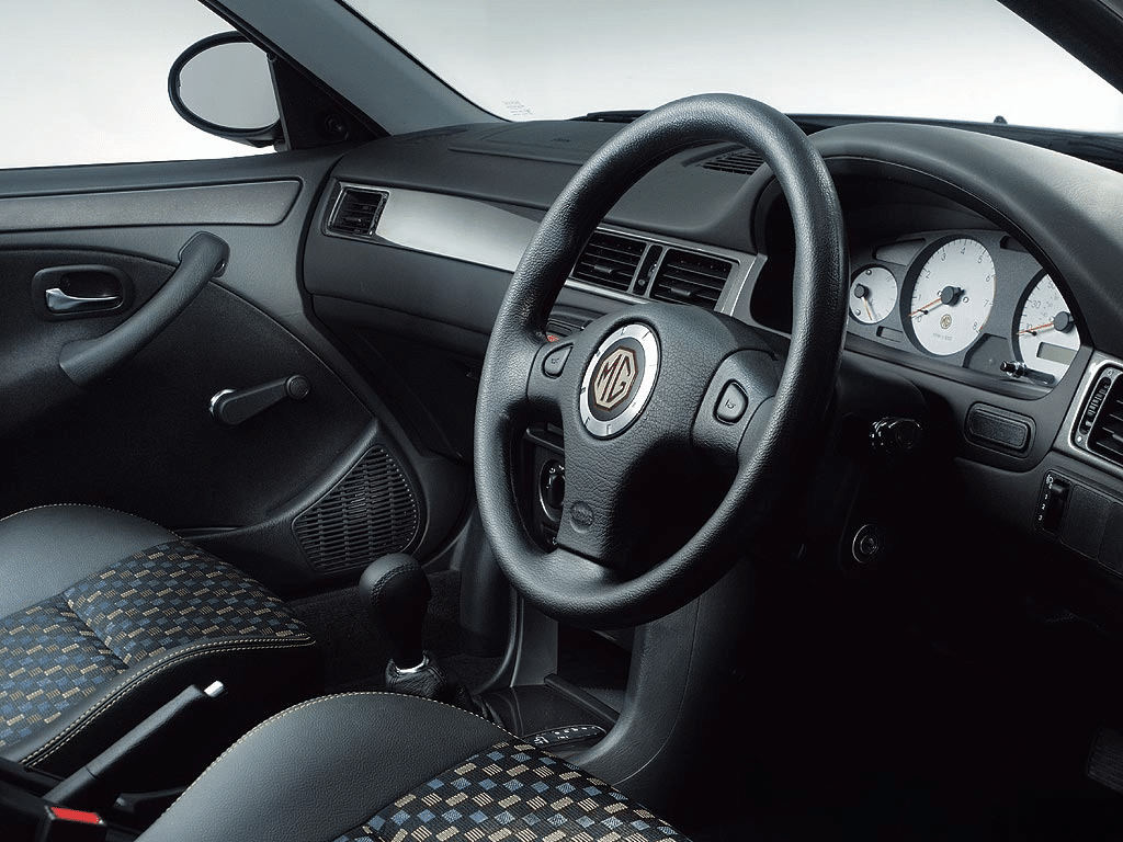 "La imagen muestra el interior de un coche, específicamente enfocándose en el lado del conductor. El volante presenta prominentemente el logotipo de MG en el centro. El tablero incluye un conjunto de indicadores con carátulas blancas, incluyendo un velocímetro, un tacómetro y otros indicadores. El diseño interior incluye una mezcla de asientos de tela negra y con patrones, controles manuales de las ventanas y una palanca de cambios. La imagen es interesante ya que destaca el diseño y las características del interior del coche, lo cual puede ser relevante para aquellos interesados en el diseño automotriz, la funcionalidad o el modelo específico de la marca MG.