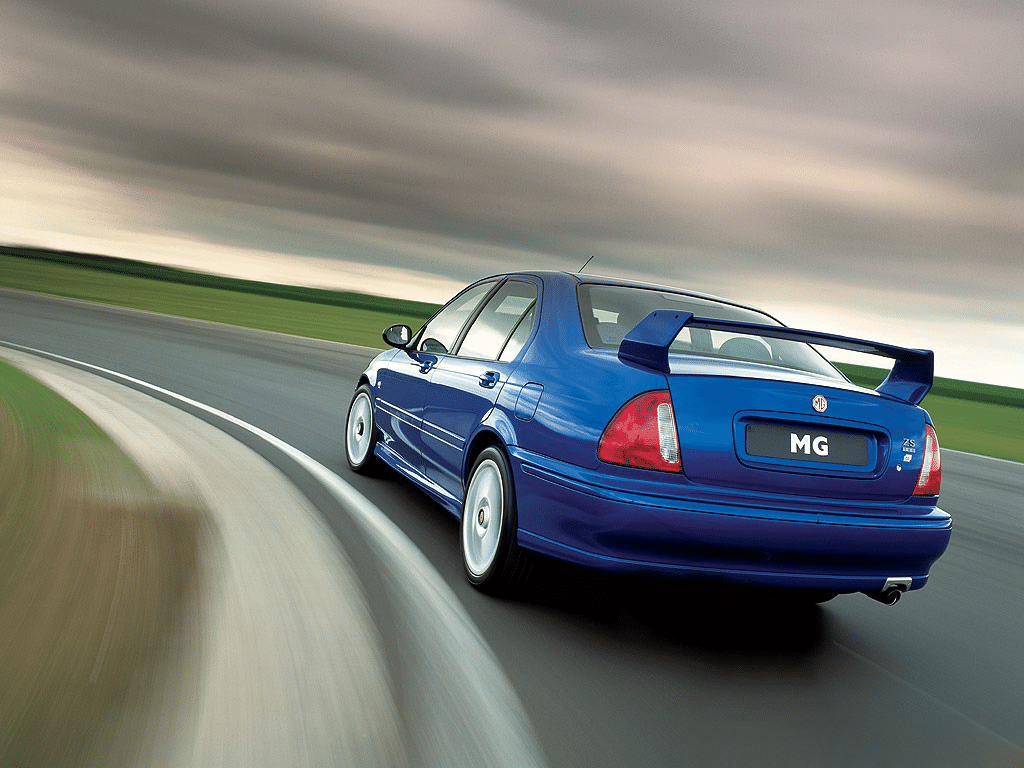"La imagen muestra un coche MG ZS 180 azul conduciendo en una pista de carreras. El coche está capturado desde la parte trasera, destacando su diseño deportivo, incluyendo un alerón trasero prominente y dos tubos de escape. El fondo está borroso, indicando movimiento y velocidad, lo que añade una sensación dinámica a la imagen. El logotipo de MG es visible en la zona de la matrícula, y la designación del modelo 'ZS 180' está en el lado derecho del maletero. Esta imagen es interesante y relevante ya que muestra las características de rendimiento y diseño del MG ZS 180, un sedán deportivo conocido por su agilidad y potencia.