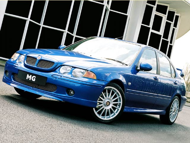 La imagen muestra un coche MG ZS 180 azul estacionado en una carretera frente a un edificio moderno con grandes ventanas. El coche tiene un diseño deportivo con una parrilla frontal prominente, llantas de aleación y una carrocería elegante. El logotipo de MG es visible en la matrícula frontal. Esta imagen es interesante porque destaca las características deportivas y elegantes del MG ZS 180, conocido por su rendimiento y diseño.