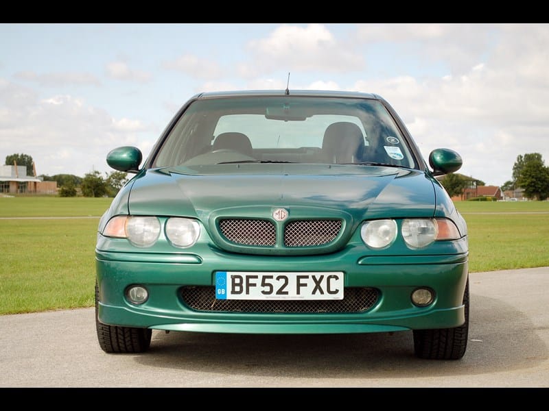 La imagen muestra la vista frontal de un coche MG ZS 180 SE verde. El coche está estacionado sobre una superficie pavimentada con un campo de césped y algunos edificios en el fondo. La matrícula dice 'BF52 FXC' y tiene una designación 'GB' en el lado izquierdo. El coche presenta una distintiva insignia de MG en la parrilla, faros dobles a cada lado y un diseño deportivo. Esta imagen es interesante porque destaca el diseño y las características del MG ZS 180 SE, un modelo conocido por su rendimiento y estilo.