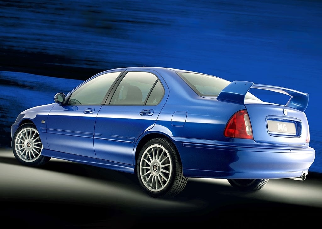 "La imagen muestra un coche MG ZS 180 azul visto desde el lado trasero izquierdo. El coche presenta un alerón trasero prominente, llantas deportivas de aleación y un diseño elegante. El fondo es un degradado de tonos azules, que complementa el color del coche. La matrícula dice 'MG'. Esta imagen es interesante ya que destaca los elementos de diseño deportivo y elegante del sedán MG, lo que puede atraer a los entusiastas de los coches o a posibles compradores que buscan un vehículo visualmente atractivo y orientado al rendimiento.