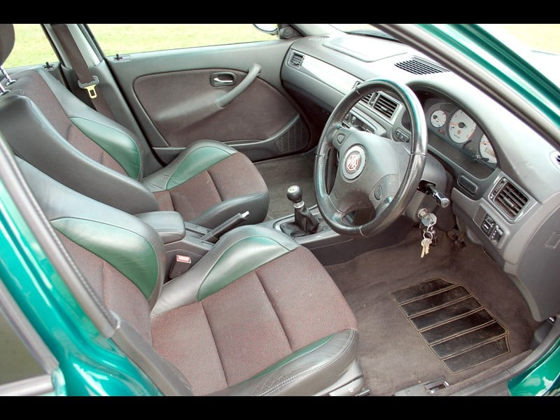 "La imagen muestra el interior de un coche MG ZS 180 SE, específicamente enfocándose en los asientos delanteros, el tablero y el área del volante. El coche tiene una configuración de volante a la derecha, lo que indica que está diseñado para países donde los vehículos circulan por el lado izquierdo de la carretera. Los asientos son una combinación de cuero y tela, con un esquema de colores en dos tonos de verde oscuro y marrón. El tablero presenta un conjunto de indicadores, una consola central con una palanca de cambios manual y varios controles. El volante tiene un logotipo en el centro, y hay llaves colgando del encendido. El suelo tiene un conjunto de pedales y una alfombrilla.