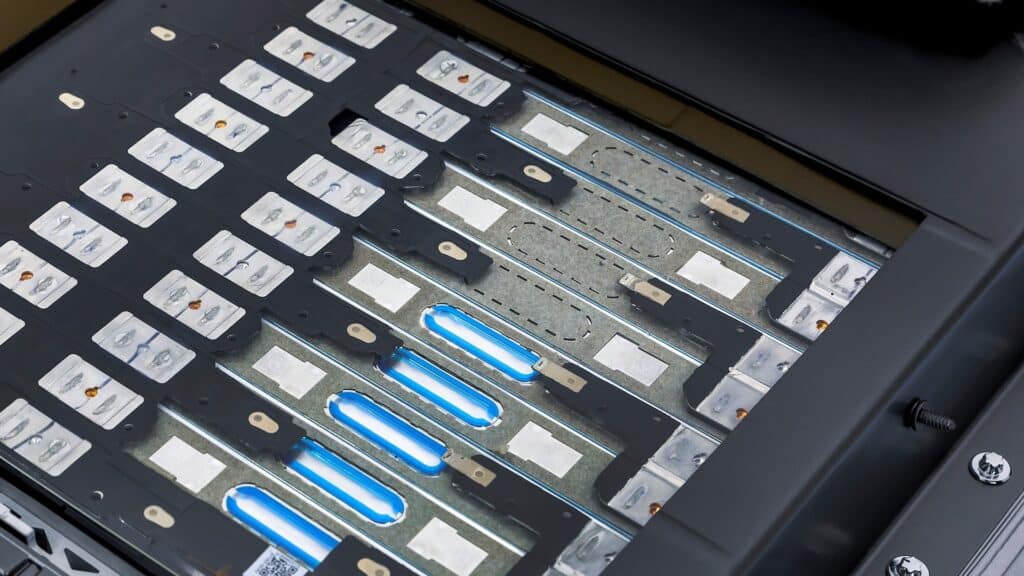 Esta imagen muestra una vista cercana de un conjunto de baterías o celdas de energía, probablemente de un vehículo eléctrico o un dispositivo similar. Se pueden observar varias celdas alineadas en paralelo, con conexiones metálicas y componentes electrónicos visibles. Algunas de las celdas están iluminadas con una luz azul, lo que podría indicar que están en funcionamiento o en un estado de carga. La imagen es relevante porque muestra la tecnología y el diseño interno de un sistema de almacenamiento de energía, lo cual es crucial para el desarrollo de dispositivos eléctricos eficientes y sostenibles.