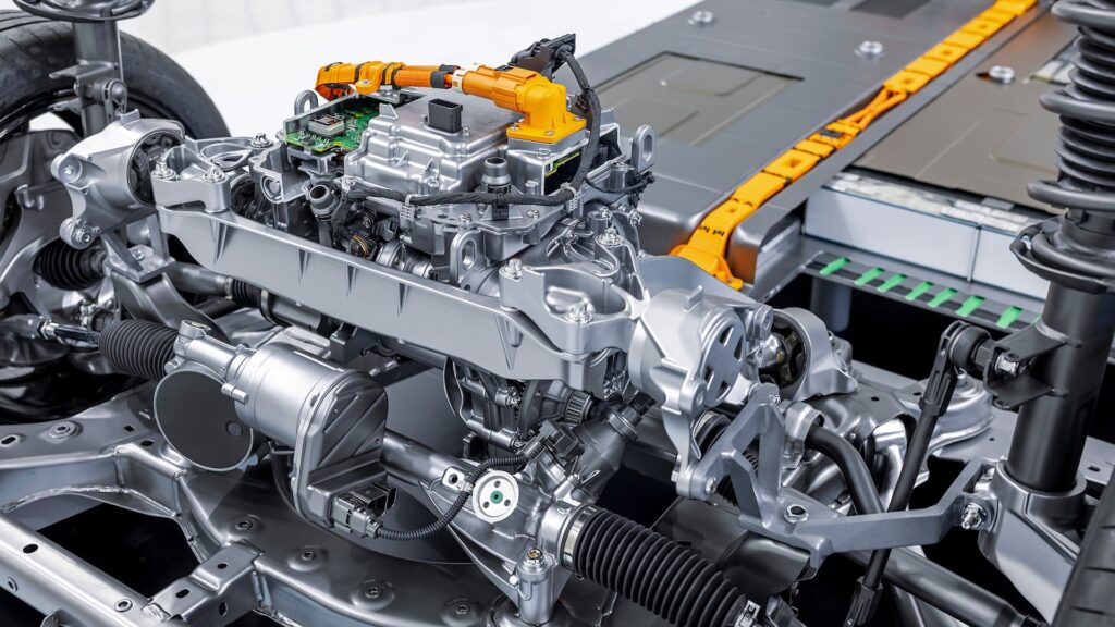 La imagen muestra una vista detallada del tren motriz y los componentes del chasis de un vehículo eléctrico. El enfoque central está en el motor eléctrico y sus componentes asociados, incluyendo la electrónica de potencia y el cableado. Los cables naranjas indican conexiones de alto voltaje, típicas en vehículos eléctricos. También se pueden ver partes del sistema de suspensión y el paquete de baterías en el fondo. Esta imagen es interesante y relevante porque proporciona una vista clara de la compleja ingeniería y tecnología involucrada en el diseño de vehículos eléctricos, destacando la integración de varios sistemas necesarios para la propulsión eléctrica.