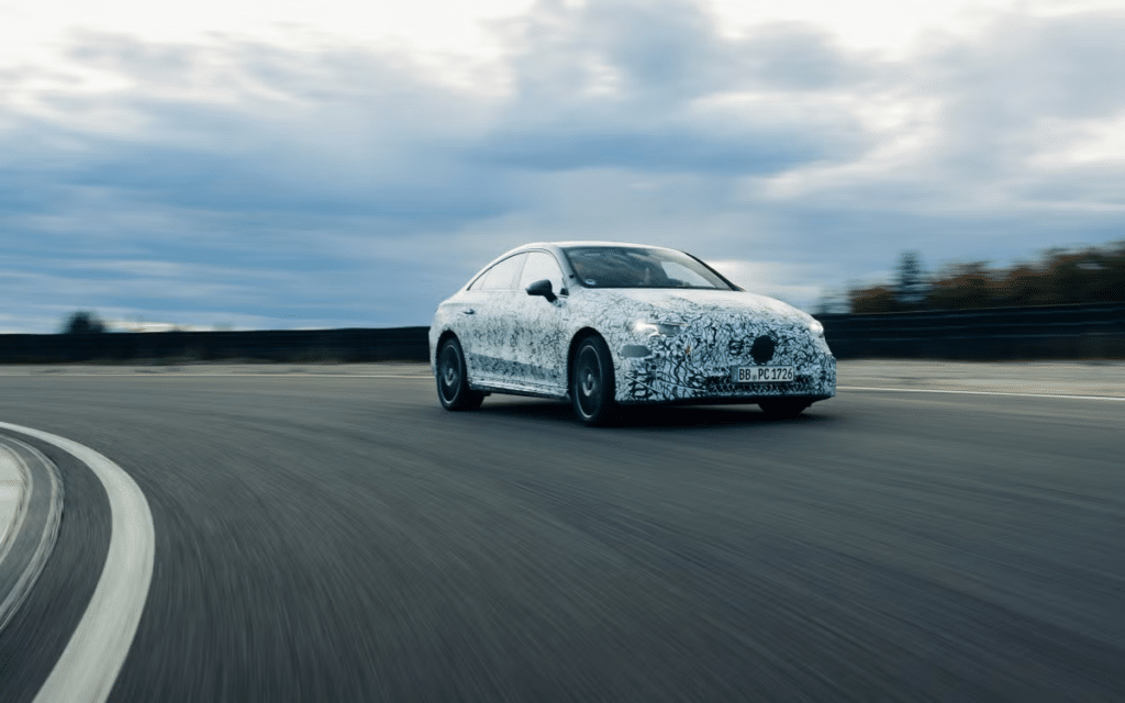 "El Mercedes CLA 2026 en movimiento en una carretera curva. El coche tiene un diseño camuflado, lo que sugiere que es un modelo de prueba o prototipo. La matrícula dice 'BB PC 1726'. El fondo muestra un cielo nublado y un paisaje borroso, indicando que el coche está en movimiento.