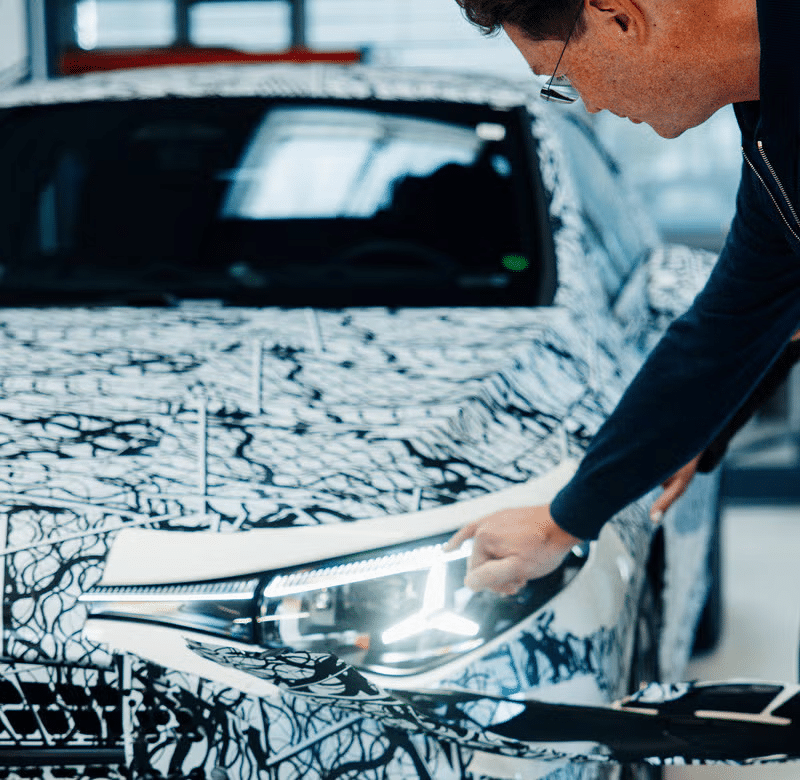 Una persona inspeccionando el faro de un coche cubierto con un camuflaje, típicamente utilizado para vehículos prototipo durante las fases de prueba. El coche está dentro de un garaje o taller bien iluminado. El faro está encendido y la persona está señalando hacia él, posiblemente verificando su funcionalidad o diseño.