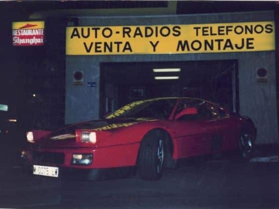 Coche deportivo rojo estacionado frente a una tienda con un cartel que dice ‘AUTO-RADIOS TELEFONOS VENTA Y MONTAJE’. El nombre de la tienda ‘Shanghai’ también es visible en el cartel. El diseño del coche sugiere que es de finales del siglo XX, posiblemente un modelo exótico o de lujo con líneas elegantes y faros emergentes. Parece ser un Ferrari 348 TB.
